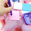 A hand uses a purple wax tool to apply clay onto pink and blue diamond painting cards on a pink textured surface.