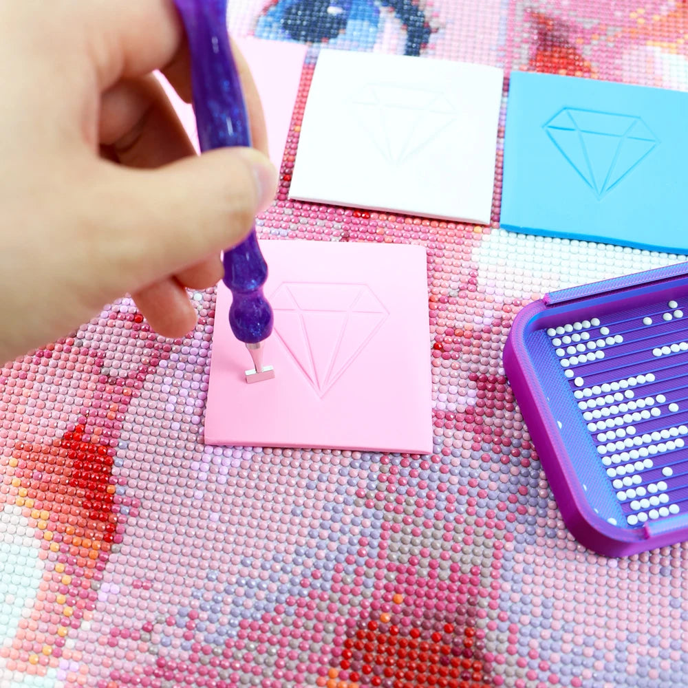 A hand uses a purple wax tool to apply clay onto pink and blue diamond painting cards on a pink textured surface.