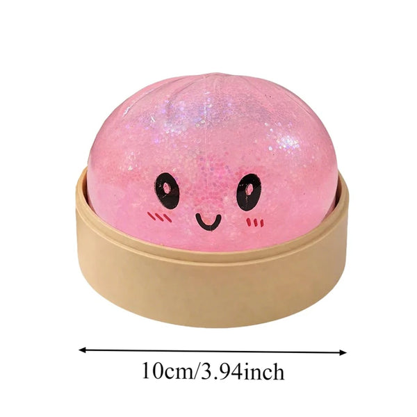 A round, pink, glittery squishy toy with simple black facial features resting in a beige container.