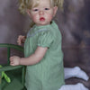 A realistic reborn baby doll with soft cloth body, dressed in a green dress and wearing white socks.