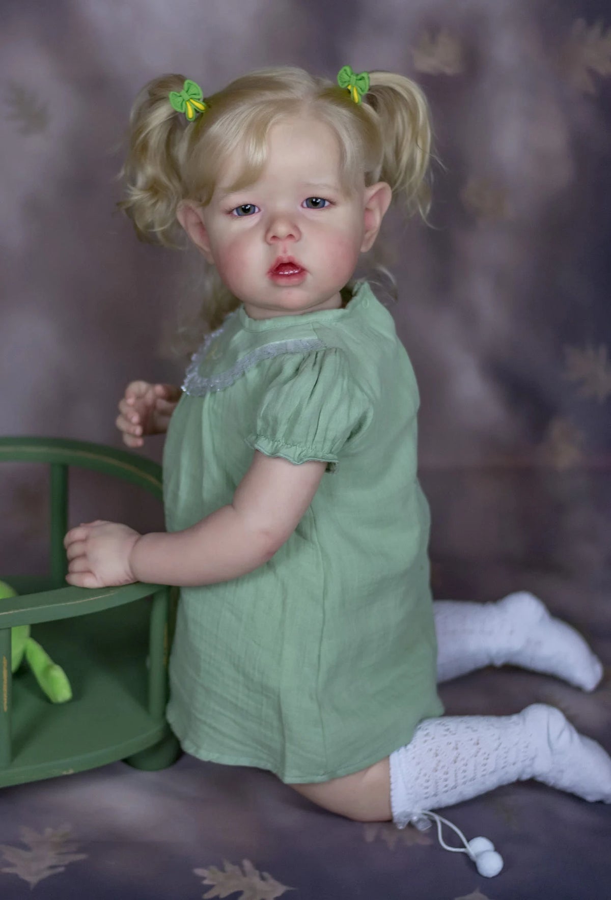 A realistic reborn baby doll with soft cloth body, dressed in a green dress and wearing white socks.