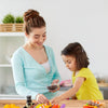 A woman is demonstrating a kids pretend play BBQ grill set with toy food on a wooden table.