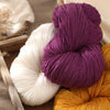 A close-up of purple, white, and orange alpaca blend yarn skeins arranged on a wooden surface.