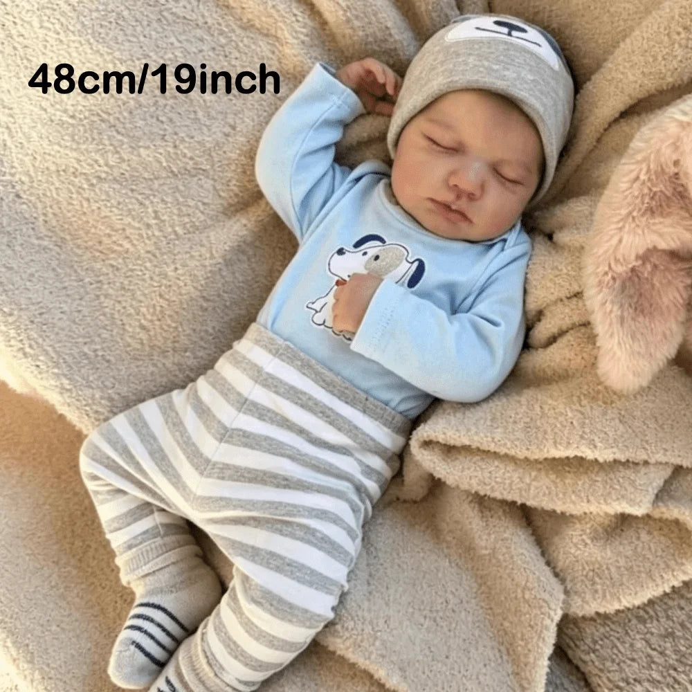 A newborn vinyl reborn baby doll wearing a light blue long-sleeved shirt and striped pants, sleeping on a beige blanket.