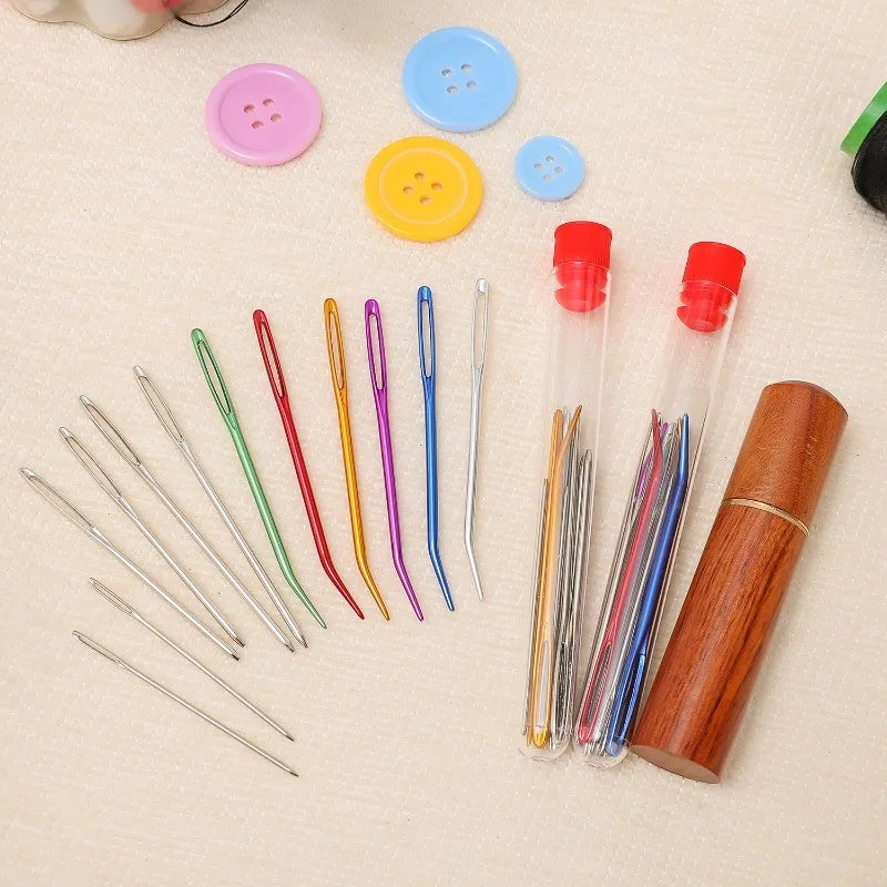 A set of large eye stainless steel sewing needles in various colors arranged next to a wooden storage tube and colorful butto