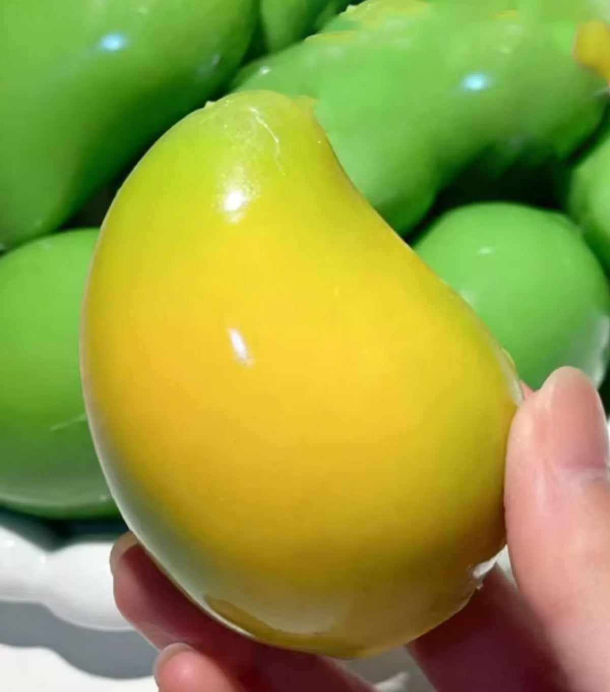 A bright yellow squishy toy shaped like a strawberry is held in a hand against a blurred background of green toys.