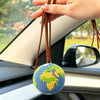 A handcrafted crochet earth keychain charm featuring a globe design, held by a hand in a car interior.