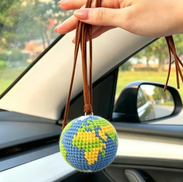 A handcrafted crochet earth keychain charm featuring a globe design, held by a hand in a car interior.