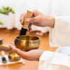 Hands are playing a golden Tibetan singing bowl with a wooden mallet and silk cushion.