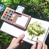 A hand holds a portable wooden watercolor paint set with a palette and a blank canvas, set against a green outdoor background