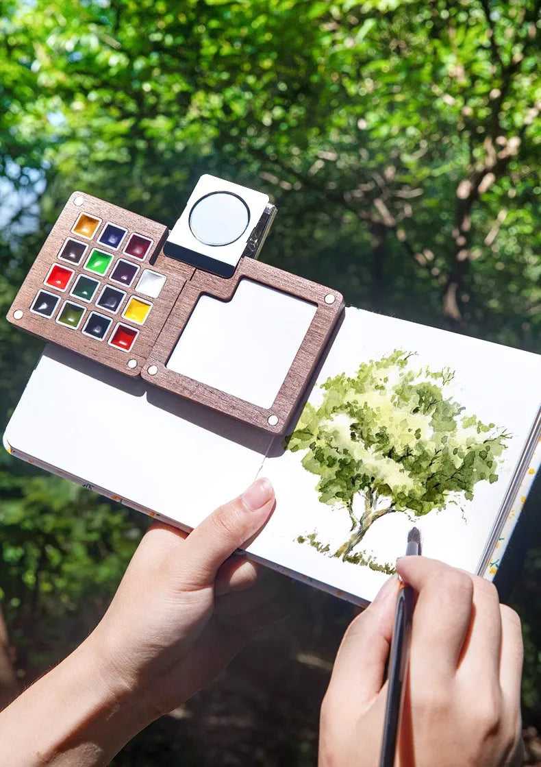 A hand holds a portable wooden watercolor paint set with a palette and a blank canvas, set against a green outdoor background
