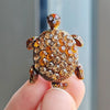 A close-up of a rhinestone turtle brooch pin with a textured, golden-brown surface held by a hand.