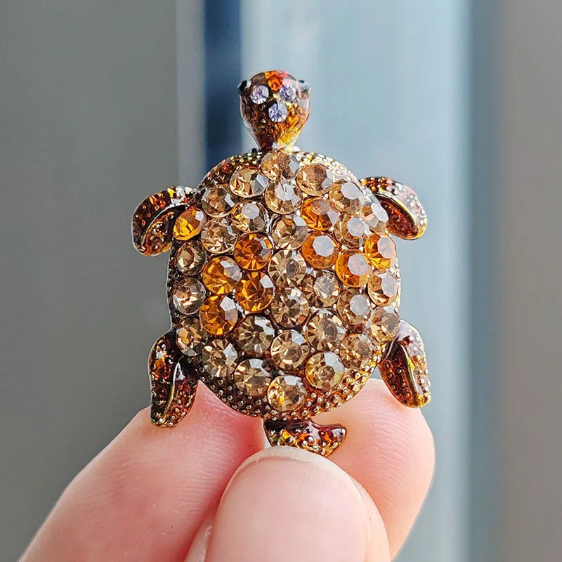 A close-up of a rhinestone turtle brooch pin with a textured, golden-brown surface held by a hand.