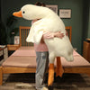 An extra-large white plush goose pillow with orange feet stands next to a wooden bed frame.