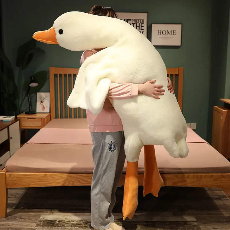 An extra-large white plush goose pillow with orange feet stands next to a wooden bed frame.