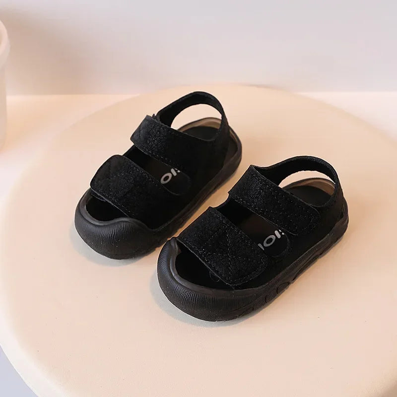 A pair of black suede toddler sandals with soft soles resting on a light surface.