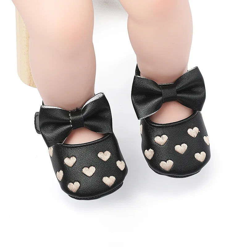 A pair of black bow-knot first walker shoes for a baby girl with a soft anti-slip sole and heart accents.