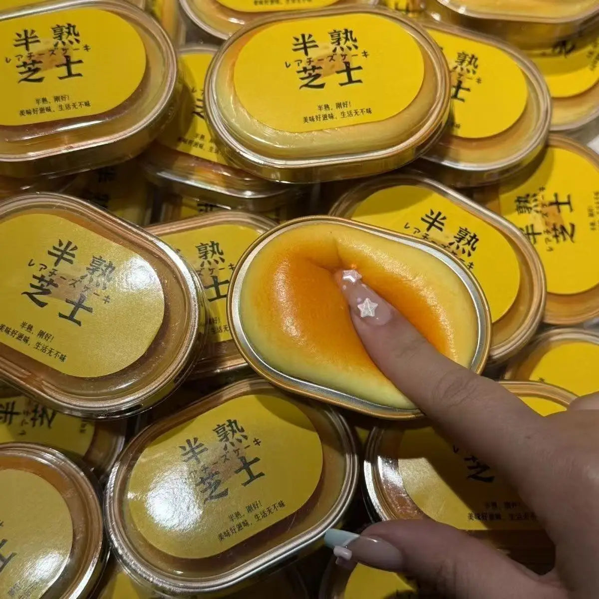 A close-up of several yellow squishy toys with Japanese text on the lid, held by a finger.