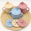 A set of nine silicone baby tableware items in pastel colors including beige, light blue, and pink, resting on a woven mat.