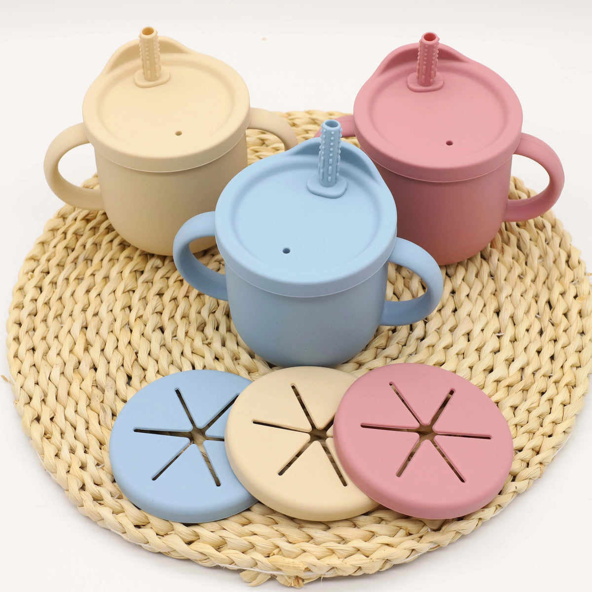 A set of nine silicone baby tableware items in pastel colors including beige, light blue, and pink, resting on a woven mat.