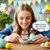 A smiling young woman holds a handmade crochet owl decoration for Easter, set against a festive background.