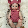 A realistic pink knitted elf hat and outfit for a reborn doll sitting in a woven basket on a fluffy white surface.