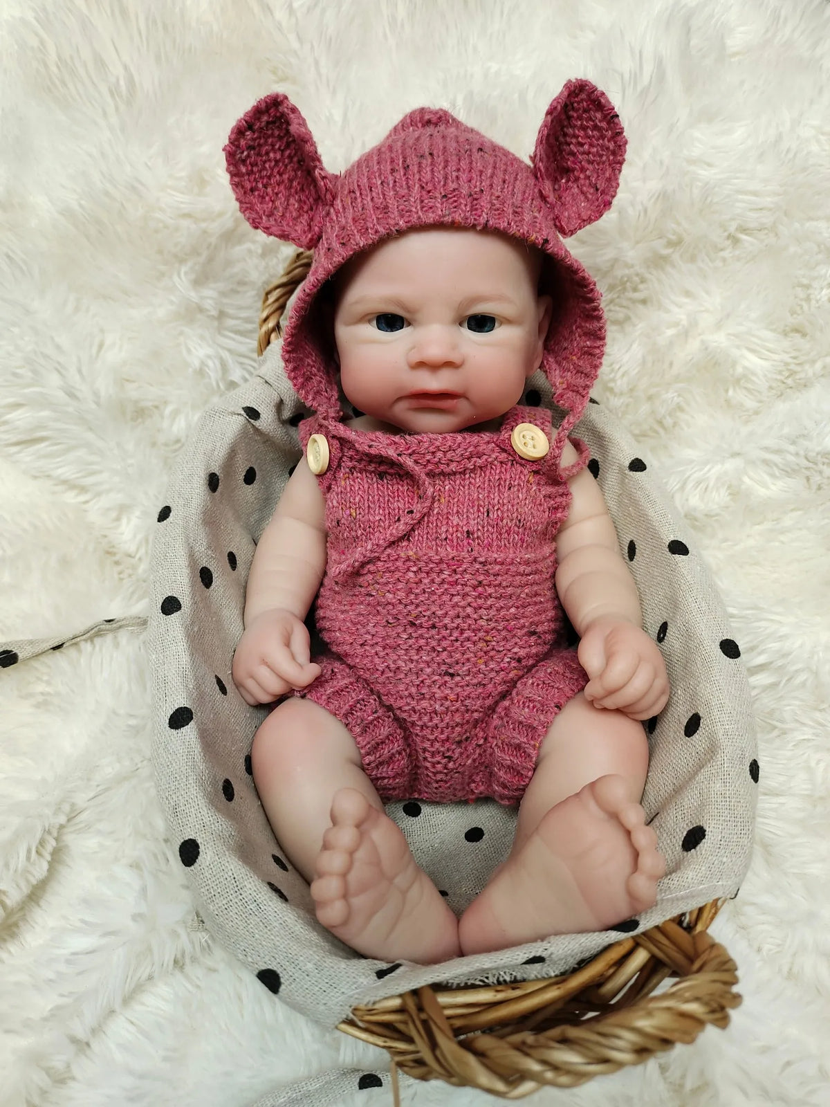 A realistic pink knitted elf hat and outfit for a reborn doll sitting in a woven basket on a fluffy white surface.
