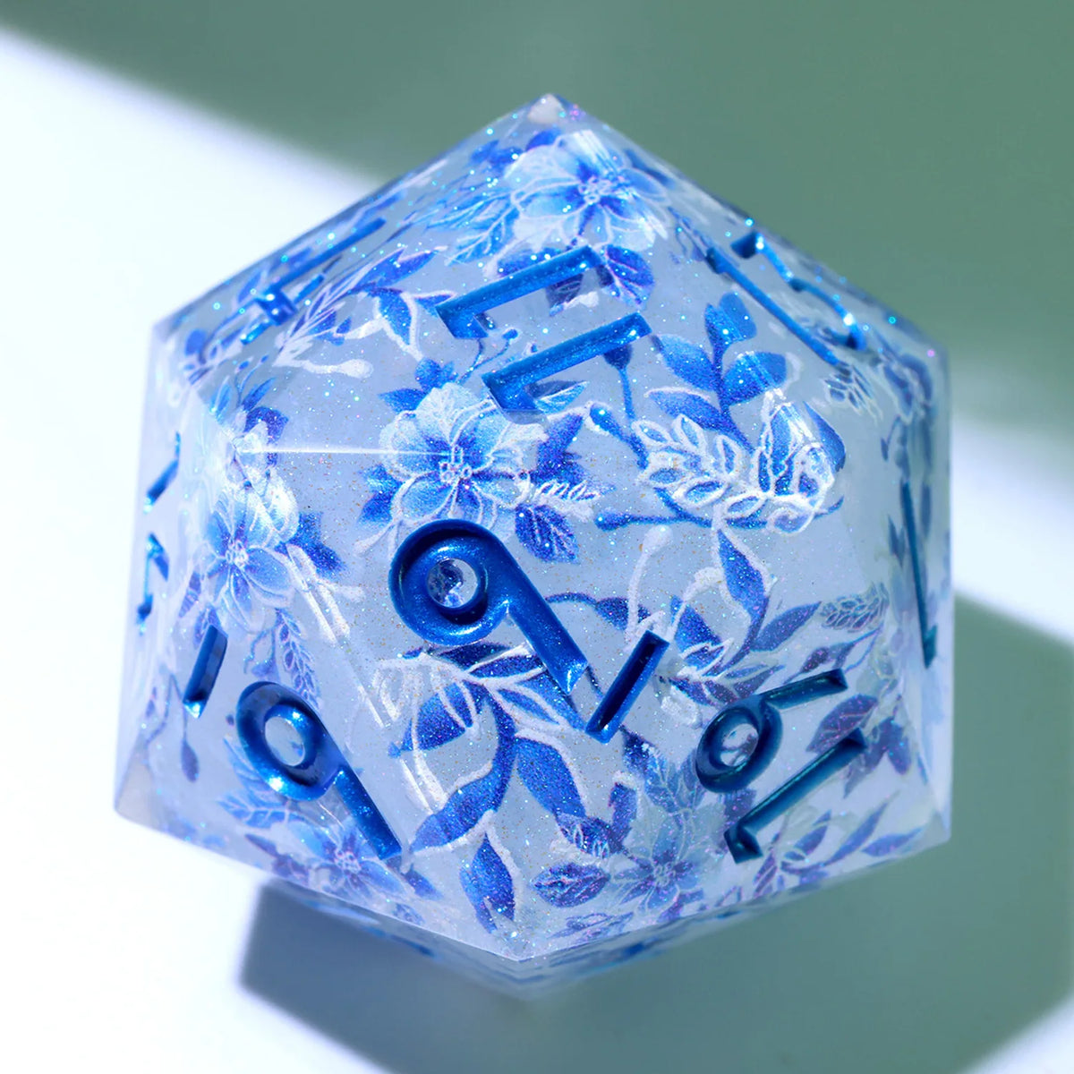 A translucent blue resin D20 die with a large, faceted shape decorated with blue and purple floral liquid swirls.