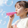 A young girl is blowing a pink smoke bubble from a handheld wand against a bright blue sky.