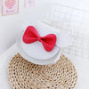 A white hard shell crossbody bag for kids decorated with mouse ears and a red bow, resting on a woven mat.