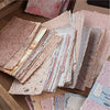 A stack of vintage scrapbook paper sheets in various shades of pink, beige, and cream inside a cardboard box.
