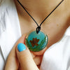 A hand holds a round, translucent teal epoxy resin pendant featuring a dried botanical element, suspended from a black cord.