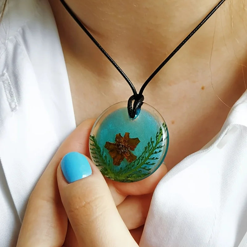 A hand holds a round, translucent teal epoxy resin pendant featuring a dried botanical element, suspended from a black cord.