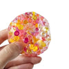 A hand holds a translucent, colorful fidget ball made of soft mochi material with various pink, yellow, and purple spheres.