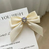 A cream colored satin bow with a clear rhinestone heart embellishment used as a hair clip.
