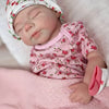 A newborn baby doll wearing a floral cap and pink shirt is sleeping on a soft pink blanket.