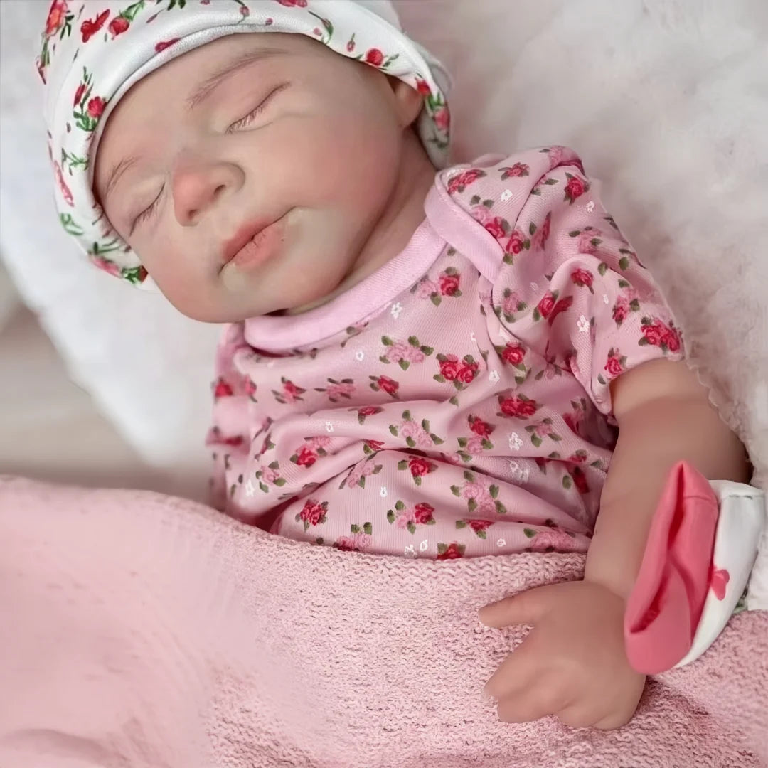 A newborn baby doll wearing a floral cap and pink shirt is sleeping on a soft pink blanket.