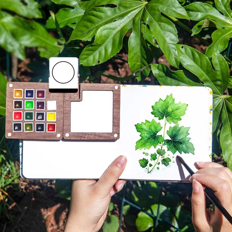 A hand holds a portable wooden watercolor paint set featuring a palette and a watercolor paper with green leaves.
