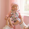 A newborn baby wearing a pink knitted hat and floral romper is being held by a person near a window.
