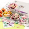 A collection of pink, green, and yellow silicone flower and leaf beads for jewelry making displayed on a white box.