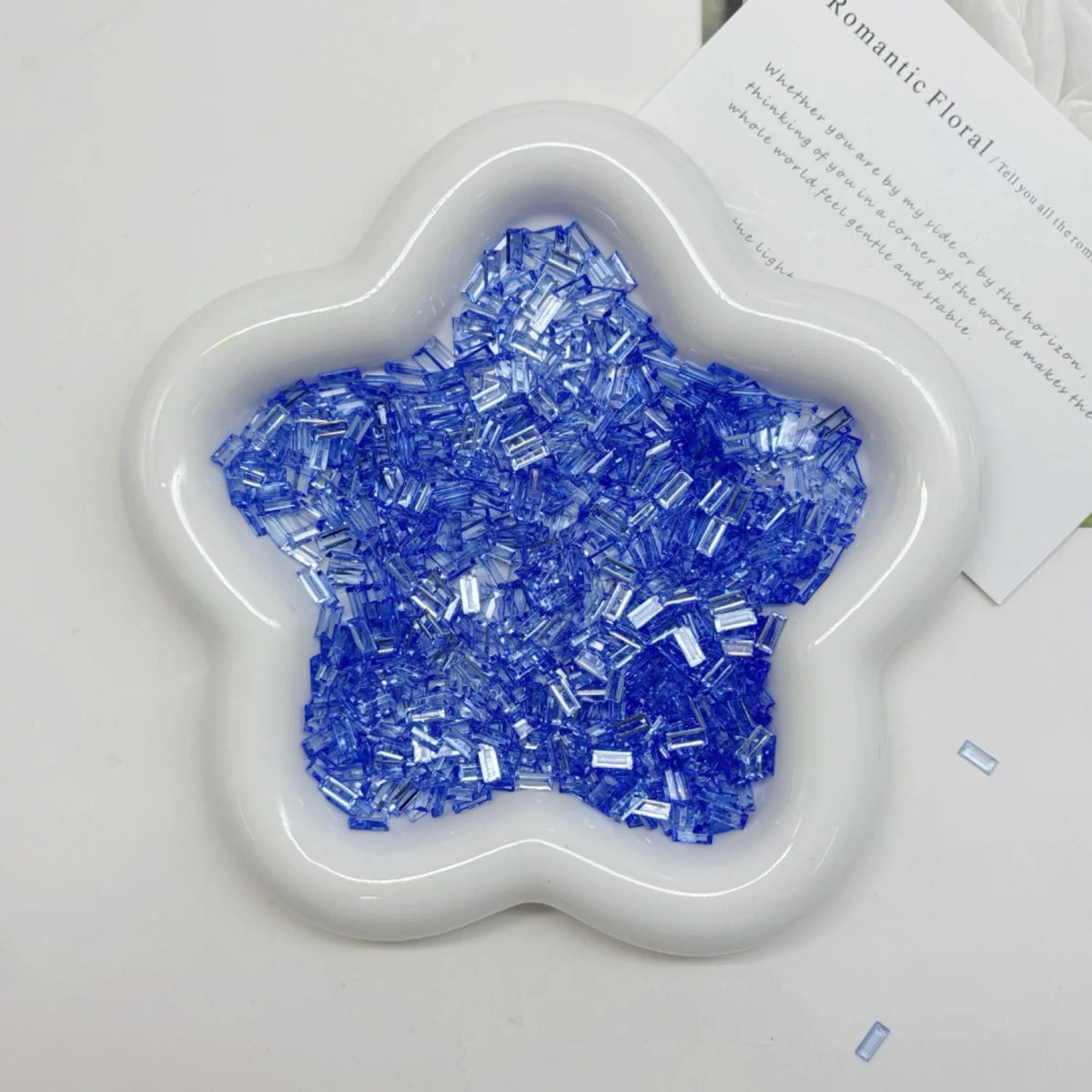A white, flower-shaped mold filled with blue iridescent acrylic ice strips for nail art and crafts.