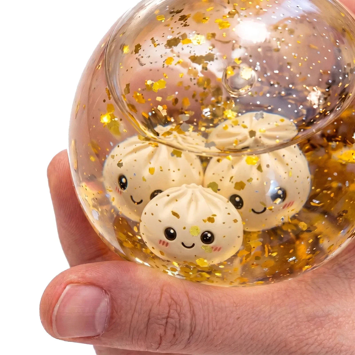 Une main tient une sphère de verre transparente remplie de paillettes dorées et de petits jouets anti-stress en forme de dump