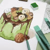 A green cake decorated with chocolate frosting and sliced apples, next to a set of painting brushes.