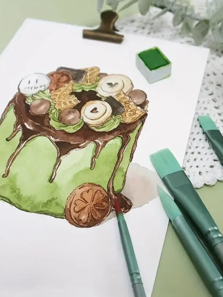 A green cake decorated with chocolate frosting and sliced apples, next to a set of painting brushes.