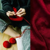 Close-up of hands knitting with red yarn against a dark sweater and denim fabric.