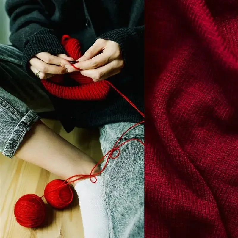Close-up of hands knitting with red yarn against a dark sweater and denim fabric.
