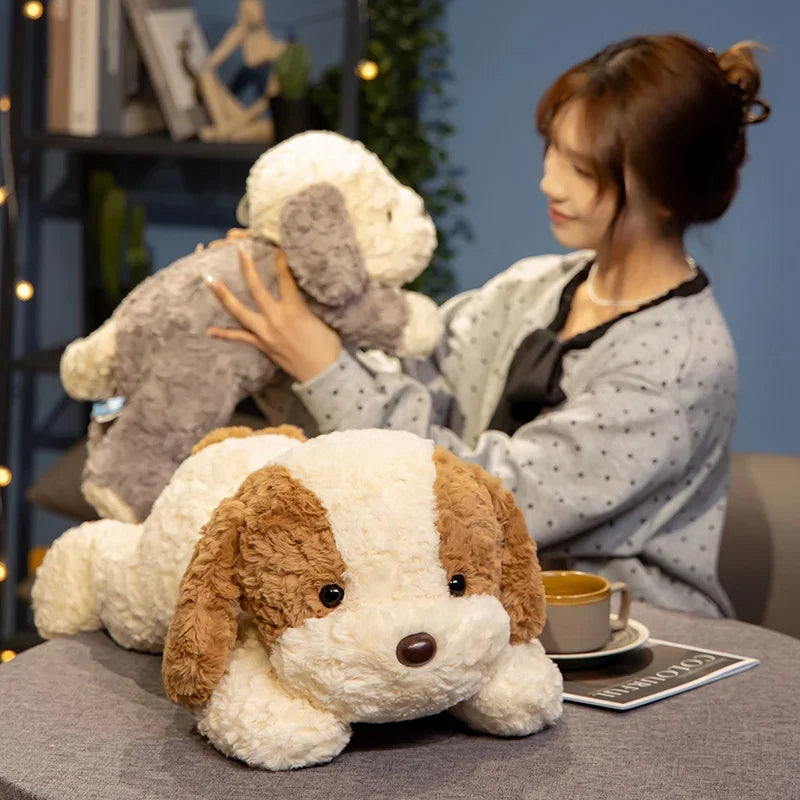 A fluffy white and brown dog plush toy lying on a table next to a person and another stuffed animal.