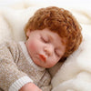 A sleeping baby doll with curly reddish-brown hair wearing a beige knit outfit, resting on a white fluffy blanket.