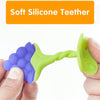A hand holds a soft silicone fruit-shaped baby teether with purple and lime green silicone parts.