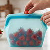 A hand is placing a strawberry into a light blue silicone storage bag filled with fresh strawberries and blueberries.